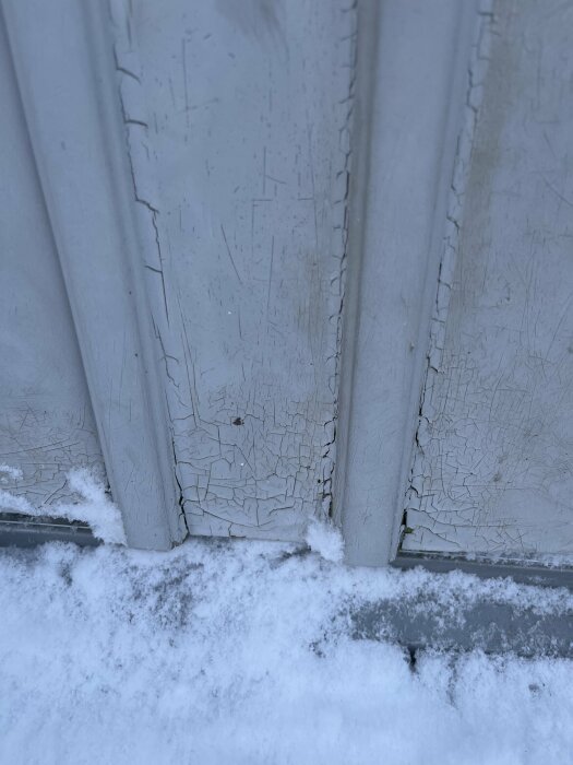 Snö mot en vägg med skalad, sprucken färg; tecken på väderpåverkan och förfall.