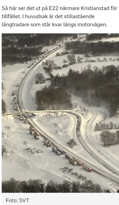 Snötäckt väg med stillastående fordon och lastbilar. Trafikstockning nära skogsområde. Bild tagen från luften.