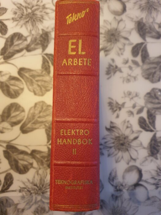 Röd bok med texten "Tekno EL ARBETE", "ELEKTRO HANDBOK II", "TEKNOGRAFISKA INSTITUTET" på ryggen.