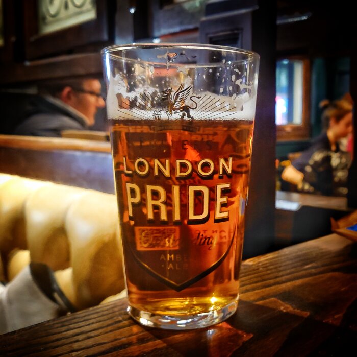 Ölglas märkt "London Pride" i förgrunden, personer i suddig bakgrund, troligtvis inne på en pub.