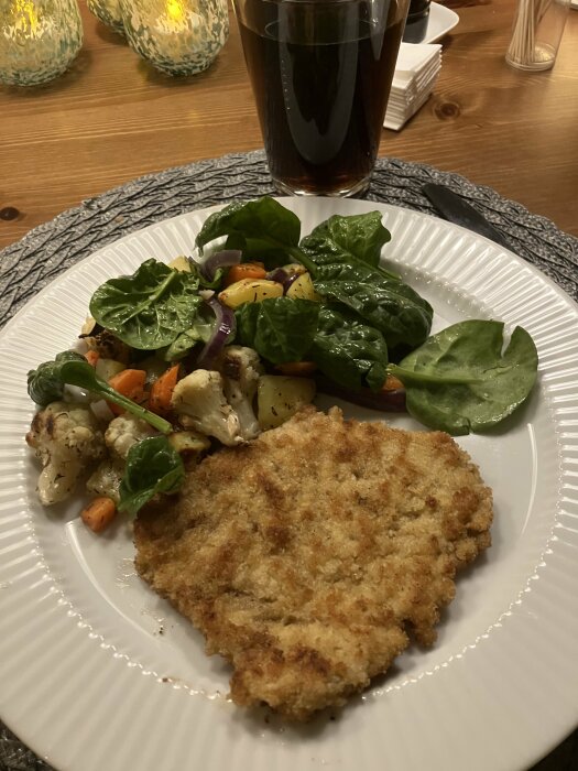 Panerad schnitzel, grönsaksblandning, spenat, på vit tallrik, mörk dryck, hemtrevlig middagsmiljö, stämningsfull belysning.