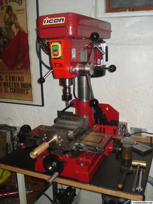 Röd bänkbordborrmaskin med flera handtag på arbetsbänk, verktyg och verkstadsutrustning runt omkring.