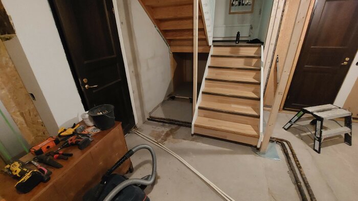 Inomhusrenovering pågår, ofärdig trätrappa, verktyg och byggmaterial synliga, dörröppningar, renoveringsstök.