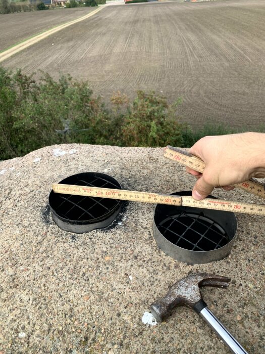 Hand mäter avstånd på betong med tumstock vid öppna runda brunnslock, hammare på marken, åker i bakgrunden.