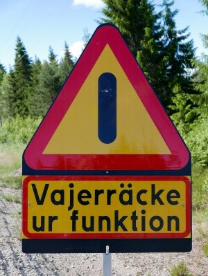 Svensk varningsskylt: röd och gul. Text "Vajerräcke ur funktion". Skog i bakgrunden.