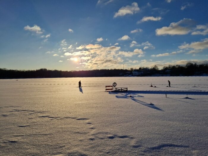 Frosten täckt sjö, personer och pulkor i solnedgången, vinterlandskap med trädstånd och moln på himlen.