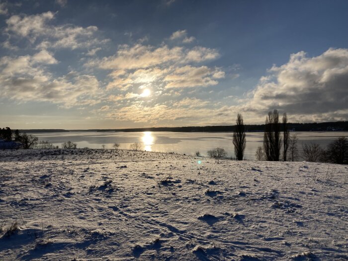 Vinterlandskap, sol över snötäckt mark och fruset vatten, moln, träd, lugnt, reflektion över is.