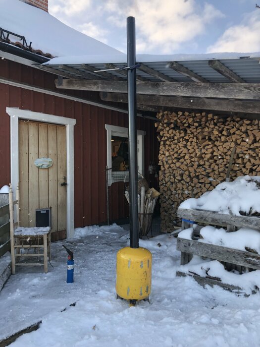 Vintrig gårdsbild med vedstapel, snö, rödmålat uthus och gul gasolflaska.