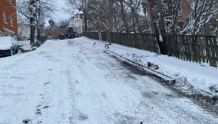 Snöig gata med traktor, snötäckta bilar, trästaket, snö på träd, övercast himmel, vinterdag.
