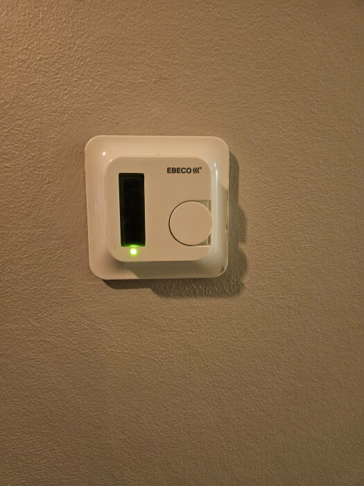 Vit termostat med display och ratt på vägg, grön lysdiod indikerar påslagen, tillverkare Ebeco.