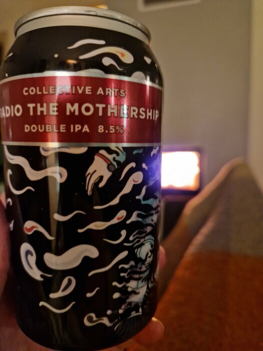 Ölburk med konstnärlig design hålls, bakgrund med mysig eld, "Radio The Mothership Double IPA 8.5%" text framträdande.