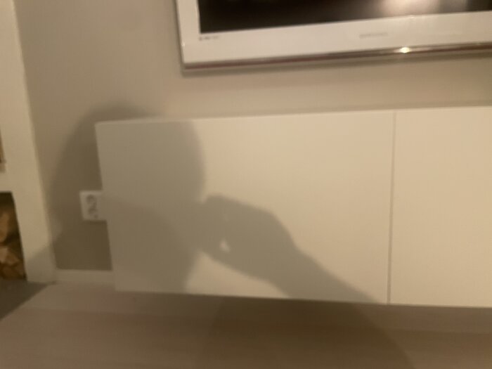 Skugga av person som håller en kamera, suddig inomhusmiljö, vägg, vit skåp, element.