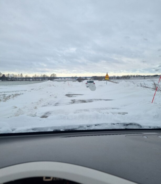 Vy från bil, snötäckt väg, avstängd vägsträcka, varningsskyltar, fält, molnig himmel, parkerad bil i fjärran.