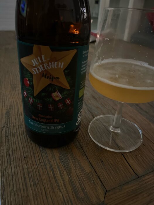 Ölflaska med etikett "Jule Stjernen Neipa" bredvid ett glas med öl, på trädgolv, inomhusbelysning.