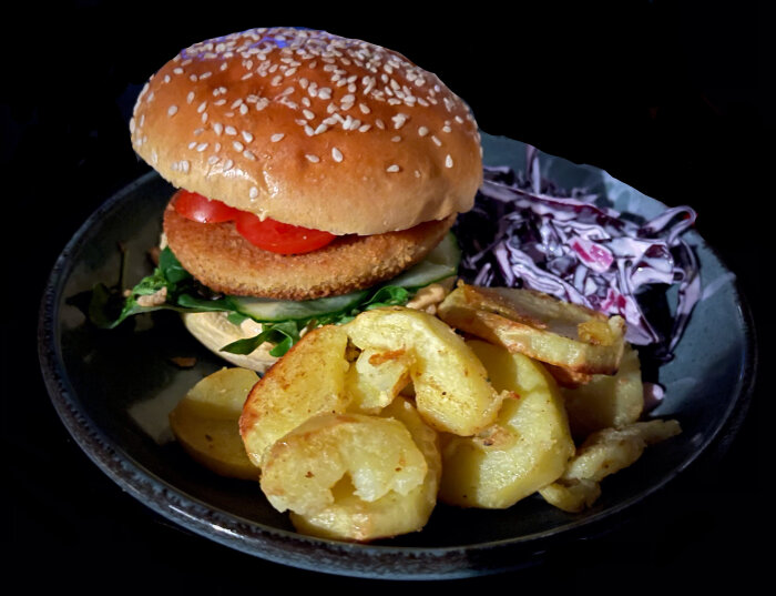 En veggieburger med sallad och tomat, rostad potatis, coleslaw på en svart tallrik.