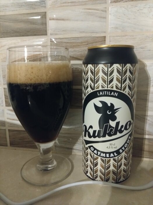 Ölglas med mörkt skum, burk med Kukko Oatmeal Stout-etikett, mörk dryck, mönstrad bakgrund.