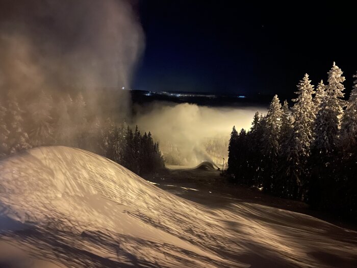 Natt, snötäckt landskap, belysta träd, konstsnöproduktion, mystisk dimma, kallt, vinteraktivitet, skidanläggning.