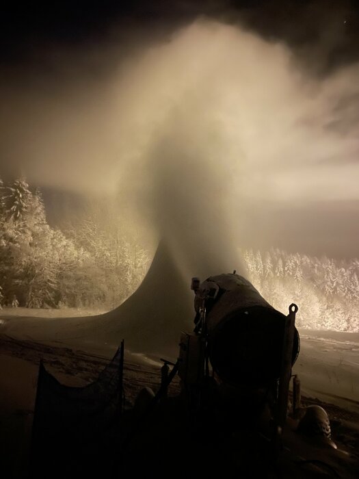 Snökanon skapar konstgjord snö nattetid; belysta träd i bakgrunden, dimmig och mystisk vinteratmosfär.