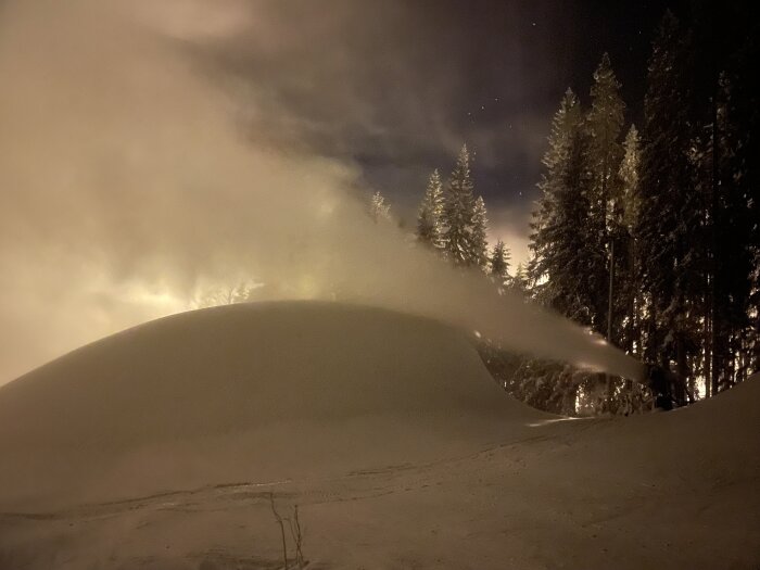 Snötäckt landskap nattetid, snökanon i drift, upplysta träd, molnigt himmel, stjärnor synliga.