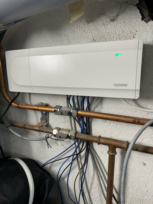 Vit Uponor-enhet på vägg, grönt ljus, kopparrör, kablar, teknisk installation, några märkningar, grå betongvägg.
