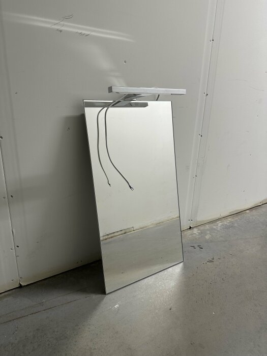 En omärkt, vit elektrisk enhet lutar mot en vägg ovanpå en spegel på ett grått golv.