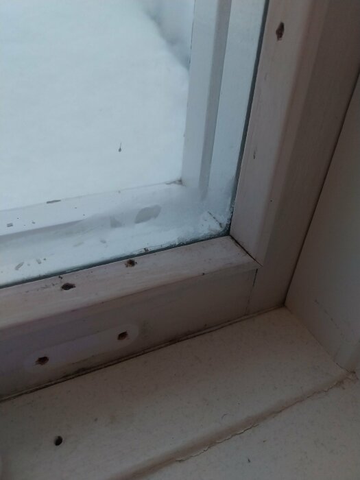 Snötäckt utsikt genom ett fönster, med synliga smutspartiklar på fönsterkarmen och fönsterblecket inomhus.