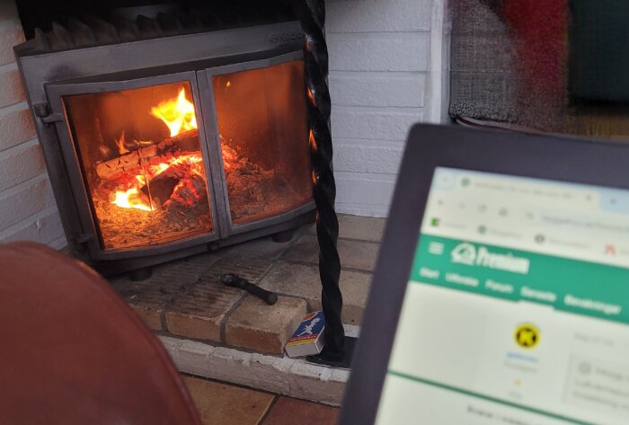 Brasvärmare med brinnande ved, aska, suddig förgrund av en laptopskärm, hemmiljö, mysigt.