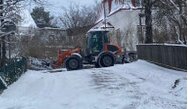En orange traktor med snöplog renar en snötäckt väg nära bebyggelse under vintern.