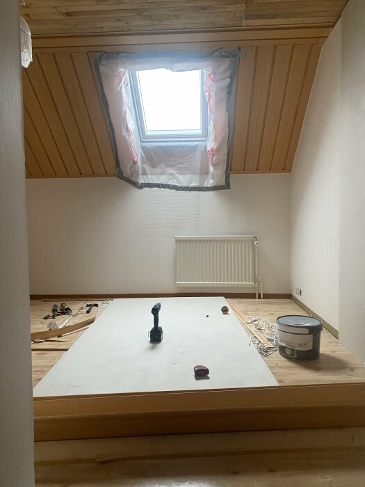 Rum under renovering, takfönster, brädor på golvet, målningsutrustning och verktyg utspridda.