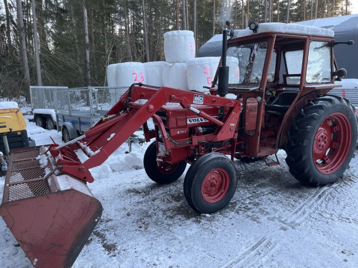 Röd Volvo traktor med frontlastare, snö, utomhus, skogsbakgrund, packade höbalar, vinterdag.