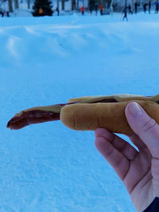 En hand håller en hotdog utomhus i ett snötäckt landskap med skidåkare i bakgrunden.