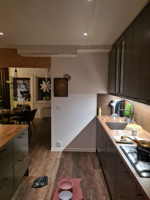 Modernt kök med mörka skåp, träbänkskiva, integrerad belysning, matplats i bakgrunden och vitt väggur.