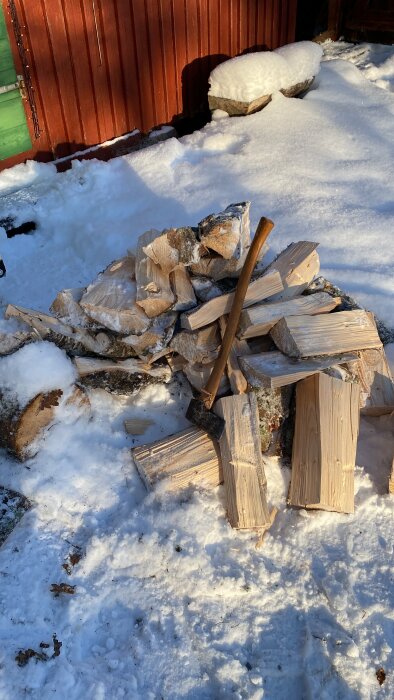 Vedkapning på vintern, yxa i snön, soligt, vedträn, röd stuga i bakgrunden.