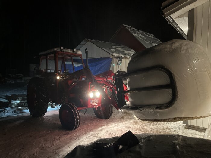 Traktor med frontlastare lyfter stor plastbal nattetid, snötäckt mark, byggnader i bakgrunden.