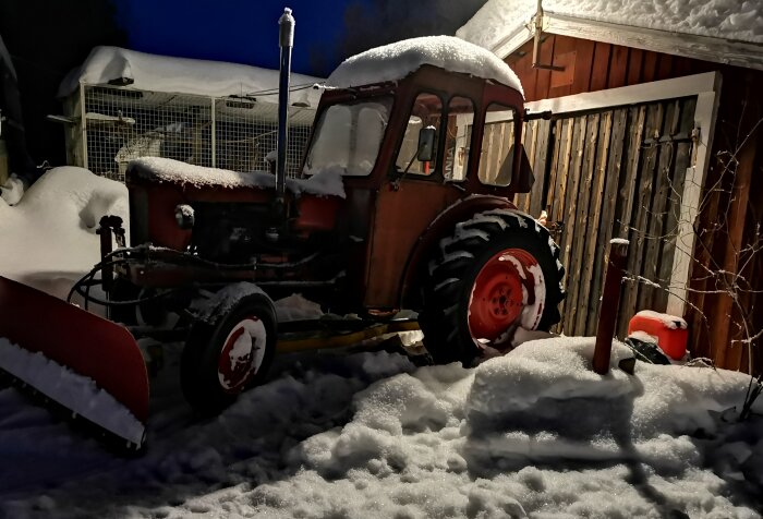 Gammal traktor med plog täckt av snö, mörk vinterkväll, snötäckt lada, lugnt och stilla.