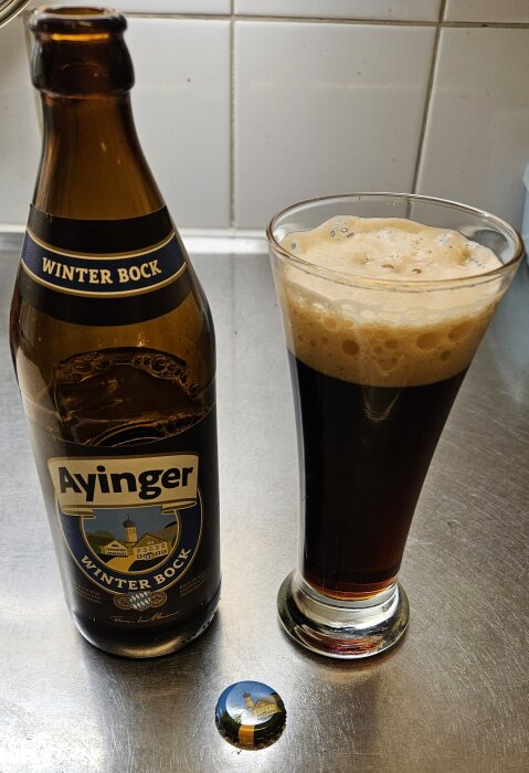 Ölflaska märkt "Ayinger Winter Bock" bredvid ett glas med mörkt öl, skumkrona, på en köksbänk.