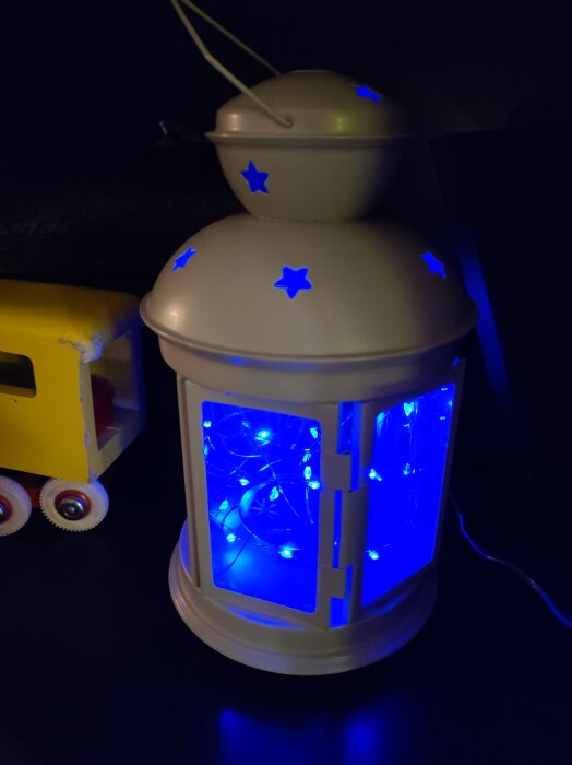 Vit lykta med blått LED-ljusslinga inuti, stjärnmönster, bredvid leksaksbuss i mörkt rum.