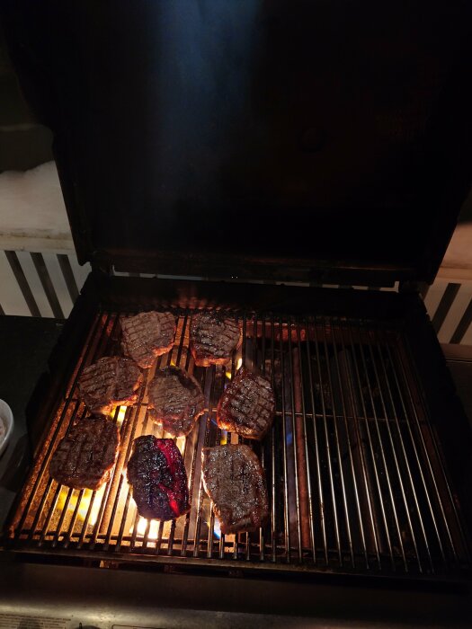 Grillad kött på en gasgrill om natten; rök syns, flammor slickar köttet.