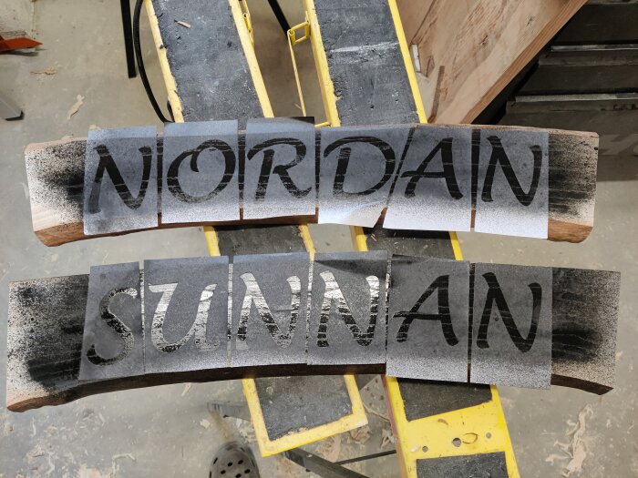 Träplankor med ord "NORDAN" och "SUNNAN" sprayade i svart på verkstadsgolv.