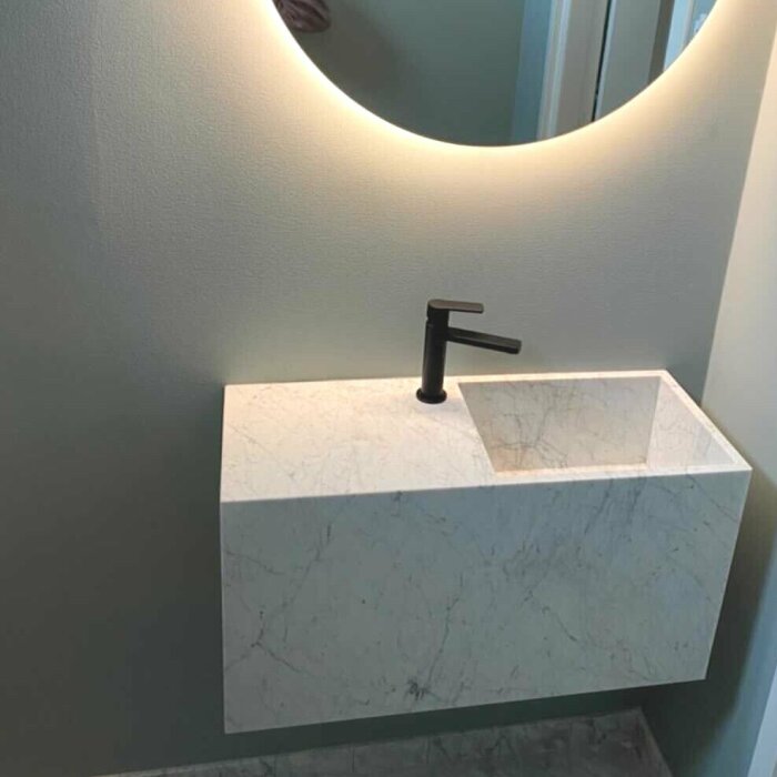 Modern handfat i marmor med svart kran, minimalistisk design, rund spegel ovanför, mjuk belysning, badrumsinredning.