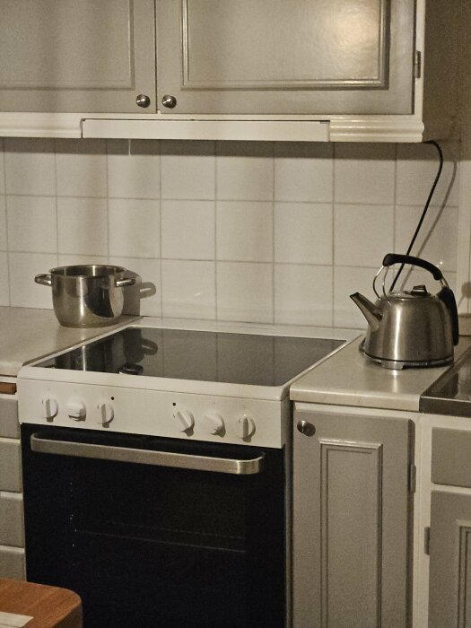Ett kök med spis, vattenkokare och kastrull, vita kakelväggar och gråa skåp.