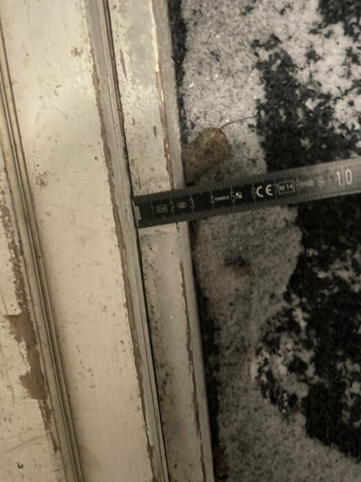 Måttband mäter glipan mellan dörr och golv; smuts, damm, och slitage synliga.