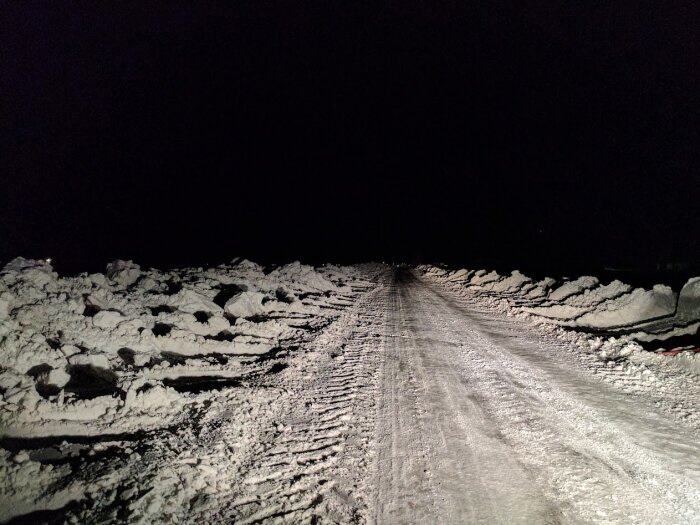 Natt, snöklädd väg, högar av plogad snö, mörker runt, bilens strålkastarljus.