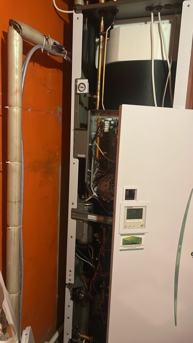 Värmesystem med öppen panel, exponerad elektronik och rörkonstruktioner, mot en orange vägg i ett teknikutrymme.