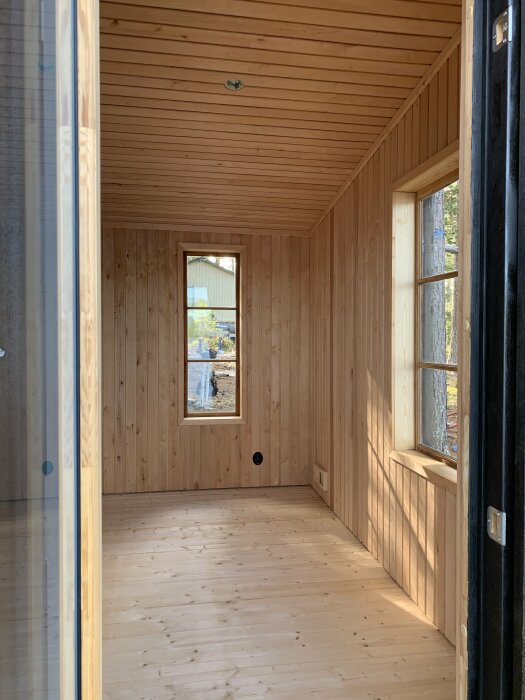 Inuti ett tomt trärum, solbelyst, med fönster, ouppvuxet trägolv, väggar och tak.