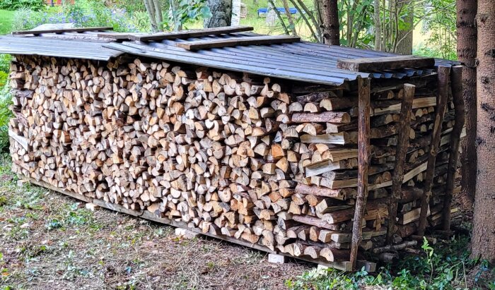 Vedtrave utomhus, stabilt staplad, skyddad med svart tak, omgiven av grönska.