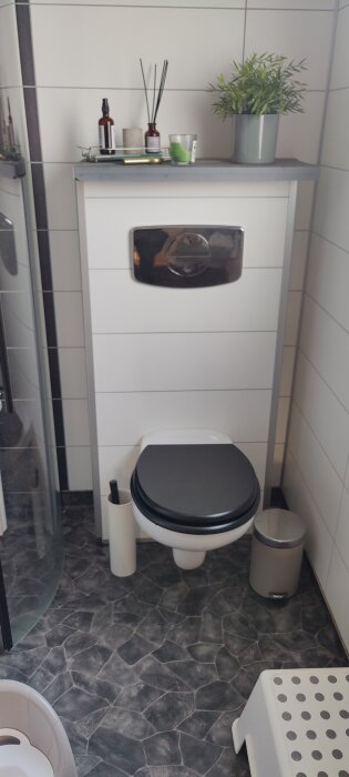 Modernt badrum med toalettstol, hyllor med växter och badrumstillbehör, pedalhink och en pall.