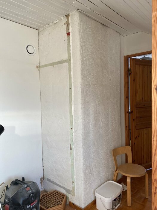 Renoveringsarbete, vitmålad vägg med avskalad yta, dammsugare, trädörr, trästol, vita takbjälkar.