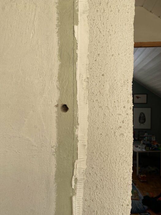 Hörn av rum med sprucken, skalad väggmålning och tapetrester, synligt hål i väggen, ojämn yta, inomhus.