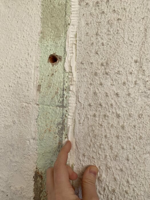 En hand pekar på skadat område på en vägg med flagnande färg och synlig struktur.